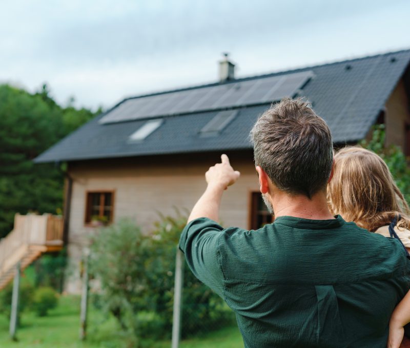 Rear view of dad holding her little girl in arms and showing at their house with solar panels.Alternative energy, saving resources and sustainable lifestyle concept.