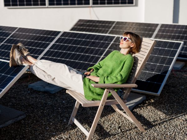 Young woman sits relaxed on a lounge chair while resting on rooftop with a solar station. Happy owner of energy-independent household. Eco-friendly and sustainable lifestyle